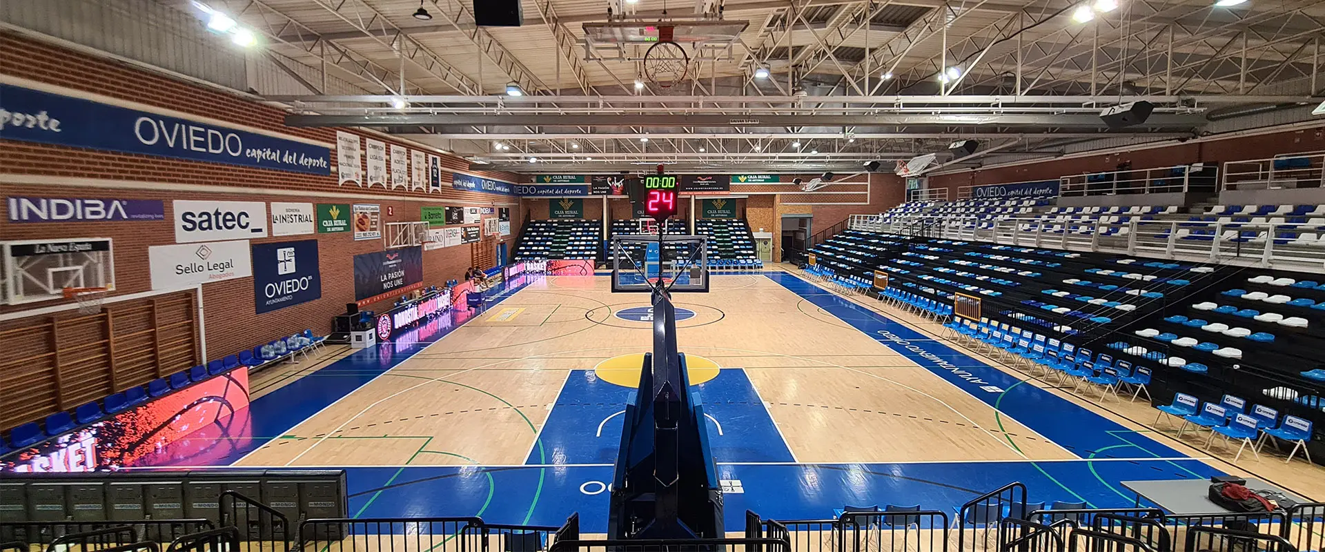 Pista polideportiva tomada desde la grada del fondo. Vista fondo, marcador y las tres gradas asientos azules y blancos