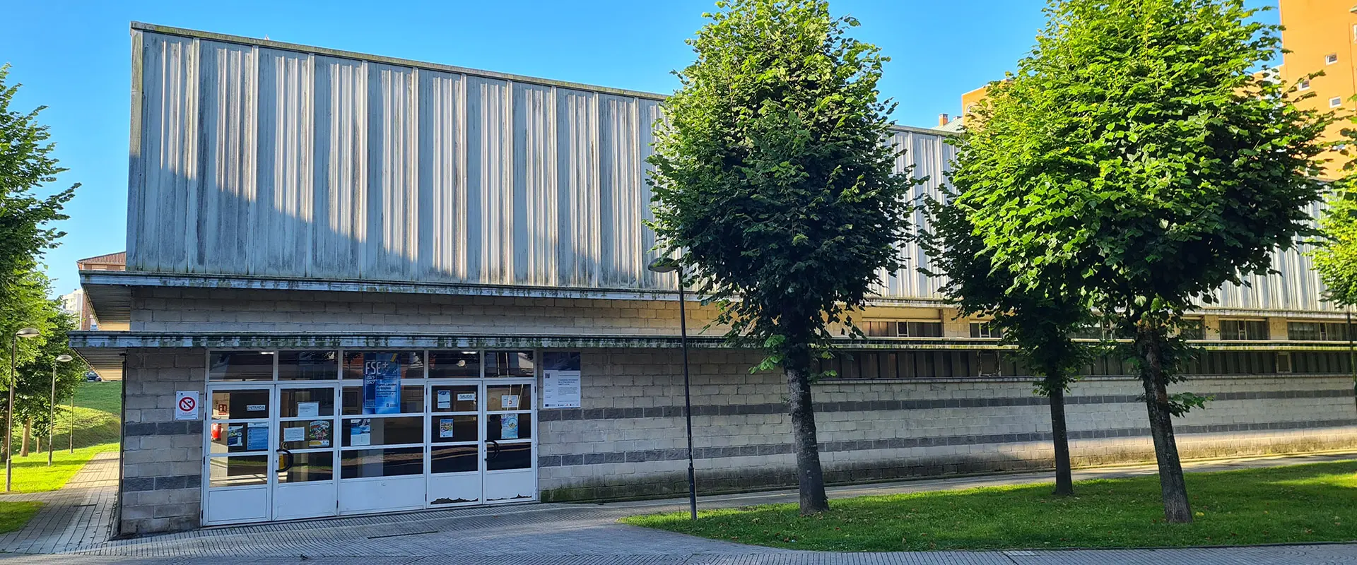 Frente de la puerta principal del Polideportivo, de metal blanco con rectángulos de cristal. En primer plano 3 árboles