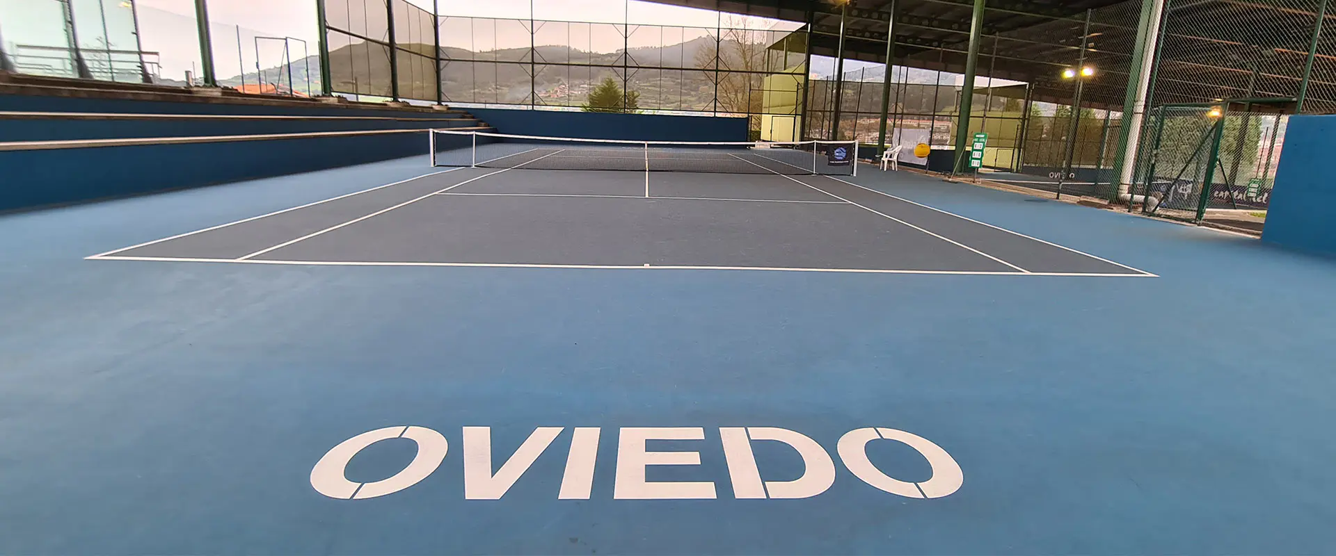 Vista a pie de pista de una cancha de tenis, con gradas, vallas de cierre y cartel de Oviedo en la pista, en primer plano