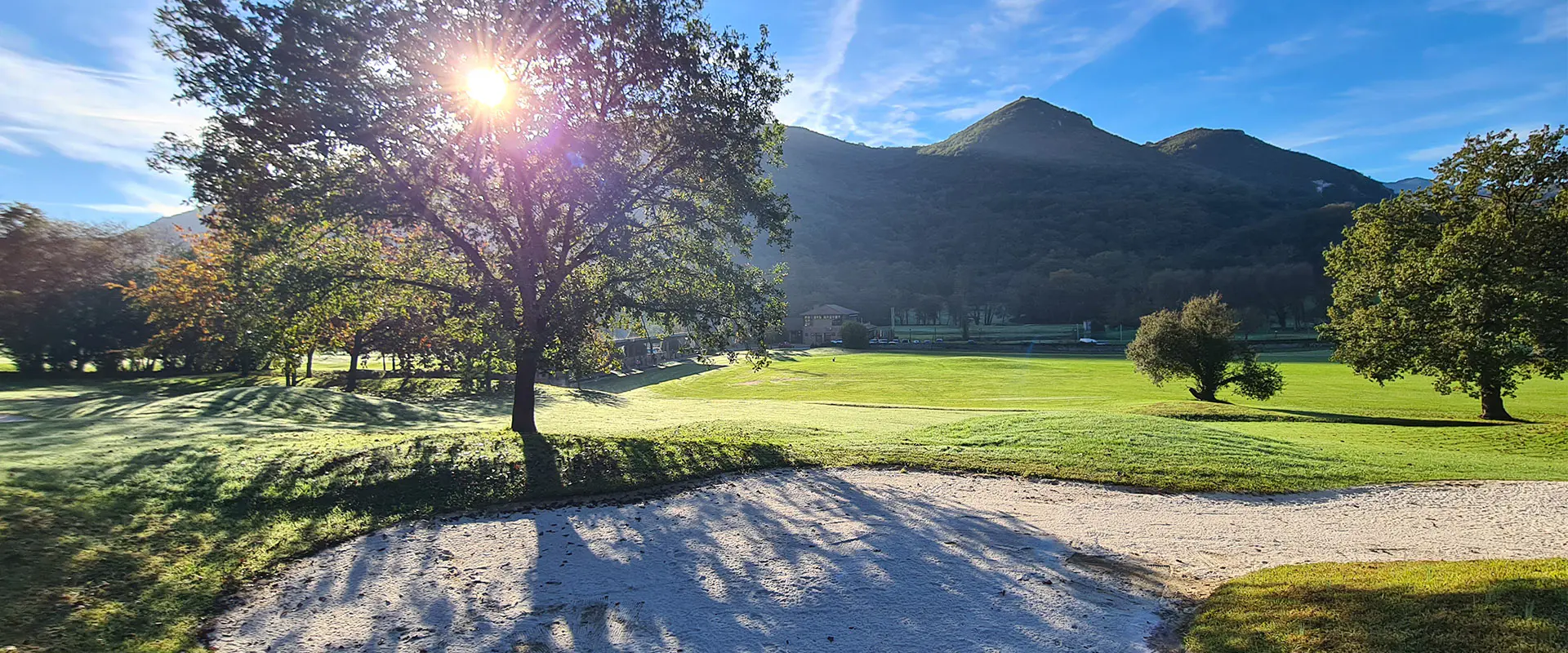 Vista de un área del campo de golf, con un búnker de arena, árboles y un fondo montañoso