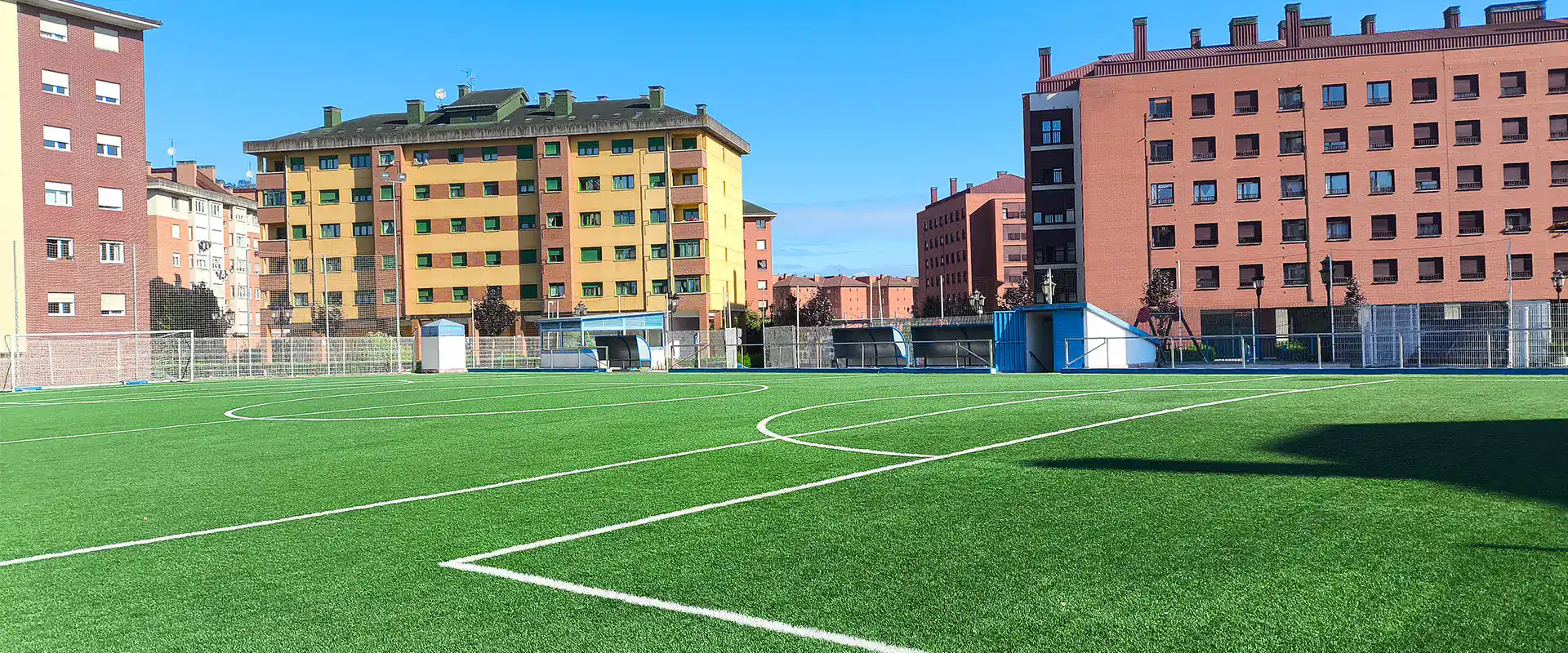 Vista lateral a pie de césped, con líneas de área en primer plano y banquillos y edificios al fondo
