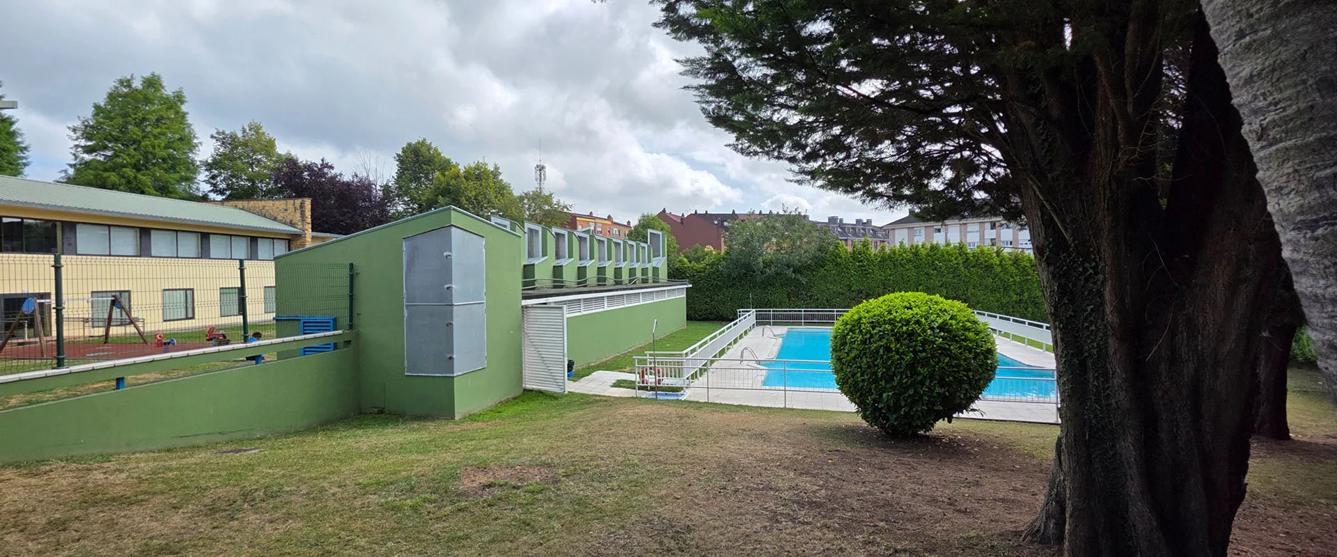 vista desde la zona de playa con árboles a la derecha, piscina al fondo y edificio de servicios a la izquierda