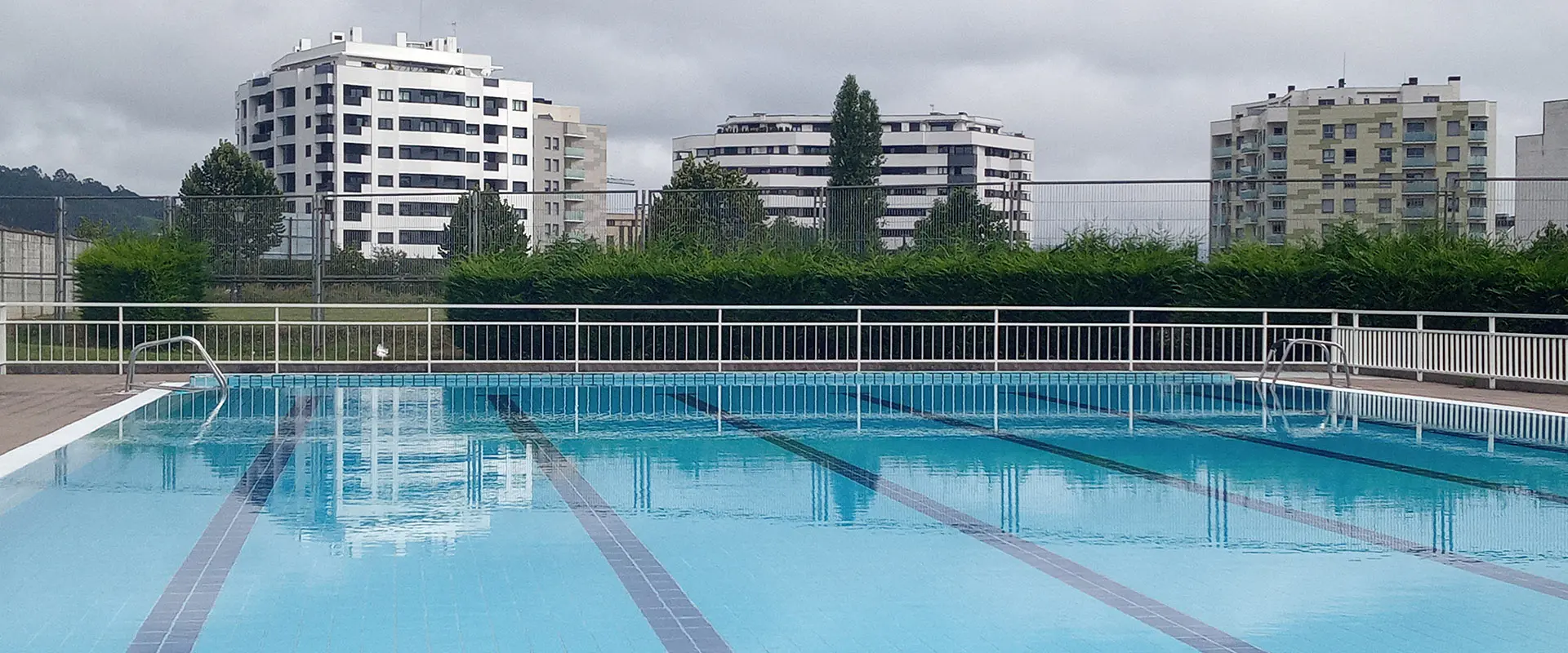 Vista lateral de la piscina principal, con las vallas perimetrales y edificios circundantes al fondo