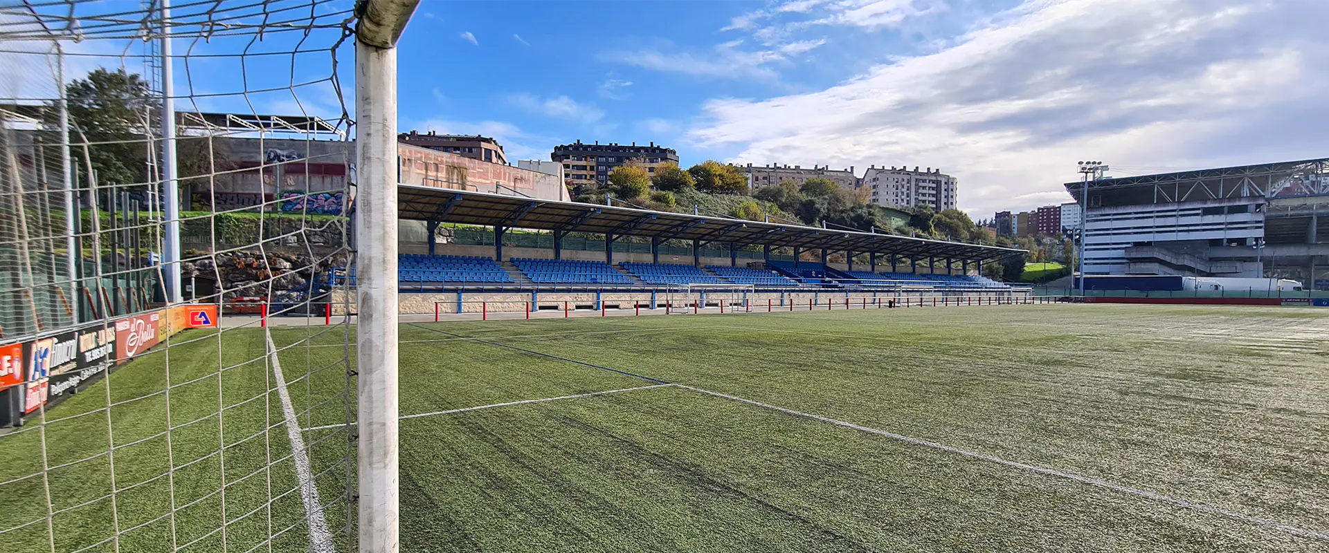 Terreno de juego en vista lateral a pie de campo. Poste y red de una de las porterías en primer plano. Gradas al fondo
