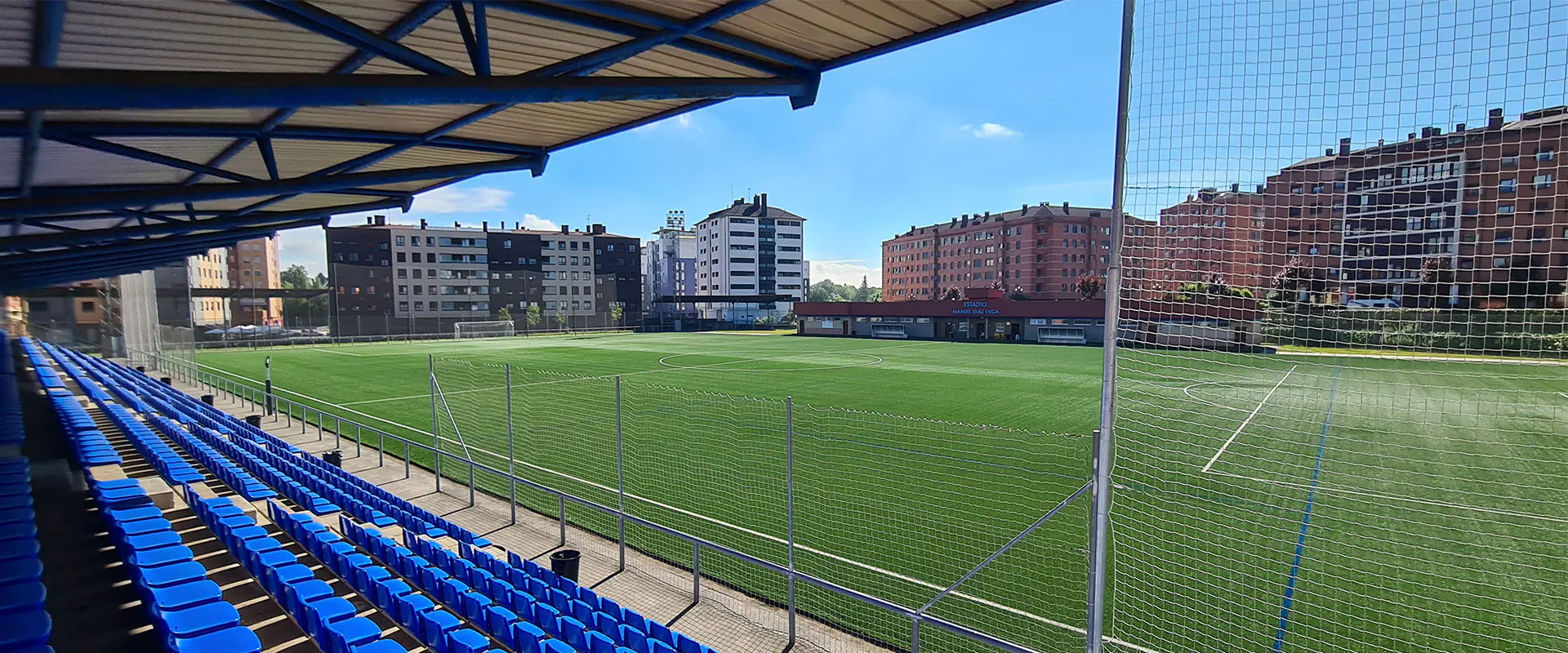 Vista lateral del campo de fútbol desde las gradas. Vallas de protección. Edificios al fondo