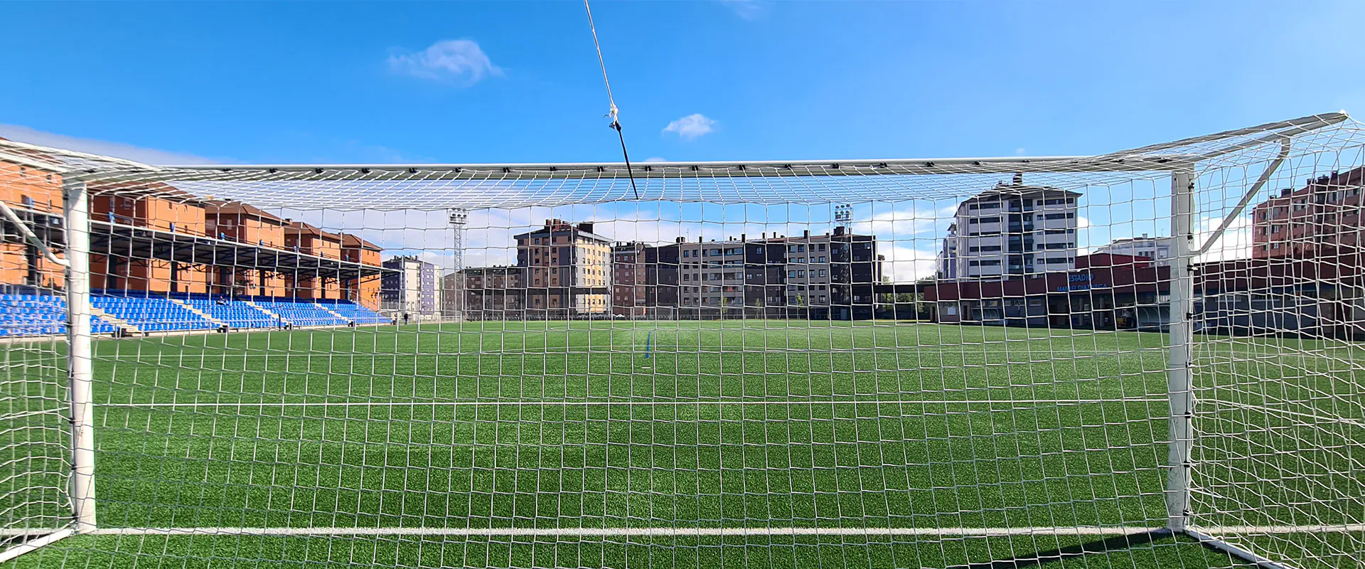 Vista frontal a pie de campo del terreno de juego, desde detrás de una de las porterías, con edificios al fondo