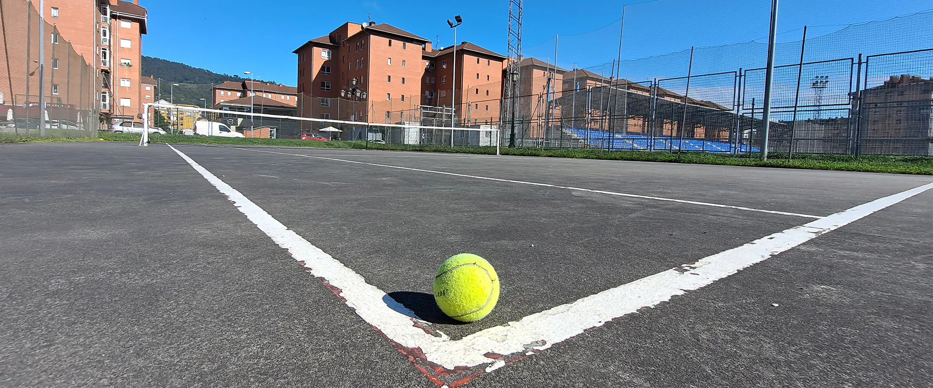 Vista lateral de una de las pistas de tenis, con una pelota en primer término y edificios de viviendas al fondo
