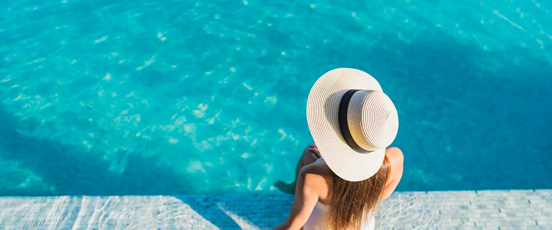 Vista parcial de una piscina con agua, con mujer rubia con sombrero sentada de espaldas en el borde