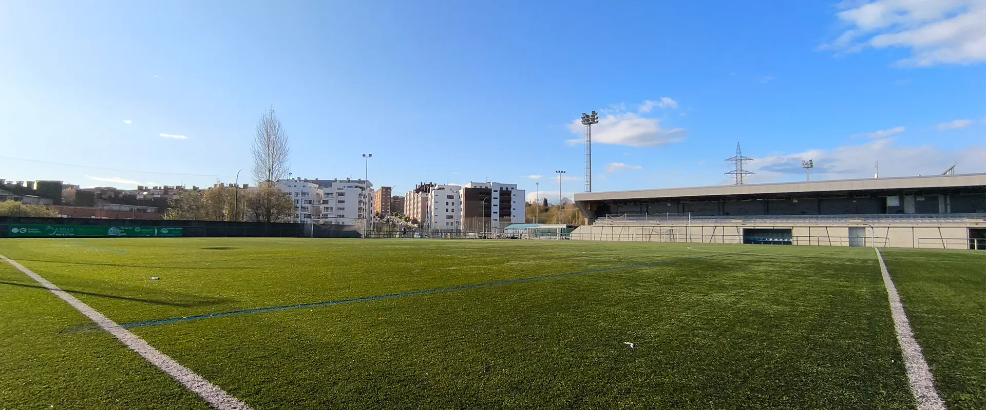 Vista a pie de campo del terreno de juego, con gradas a un lado y torre de iluminación y edificios cercanos al fondo