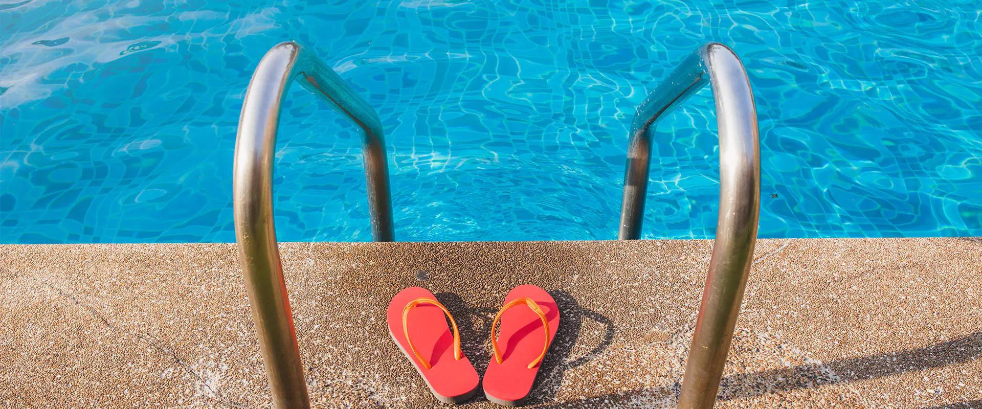 Vista parcial de una piscina con agua, con detalle de las barandillas de acceso y dos sandalias rojas entre ellas