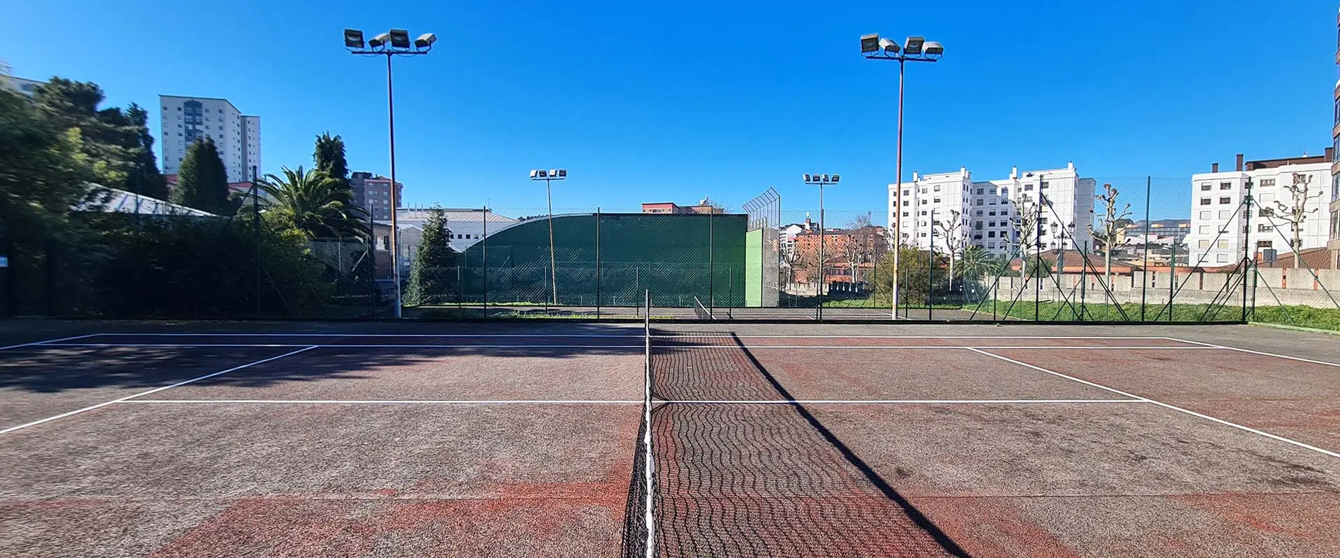 Vista lateral de una pista de tenis, con la red central, vallas, torres de iluminación y, al fondo, la pared de un frontón