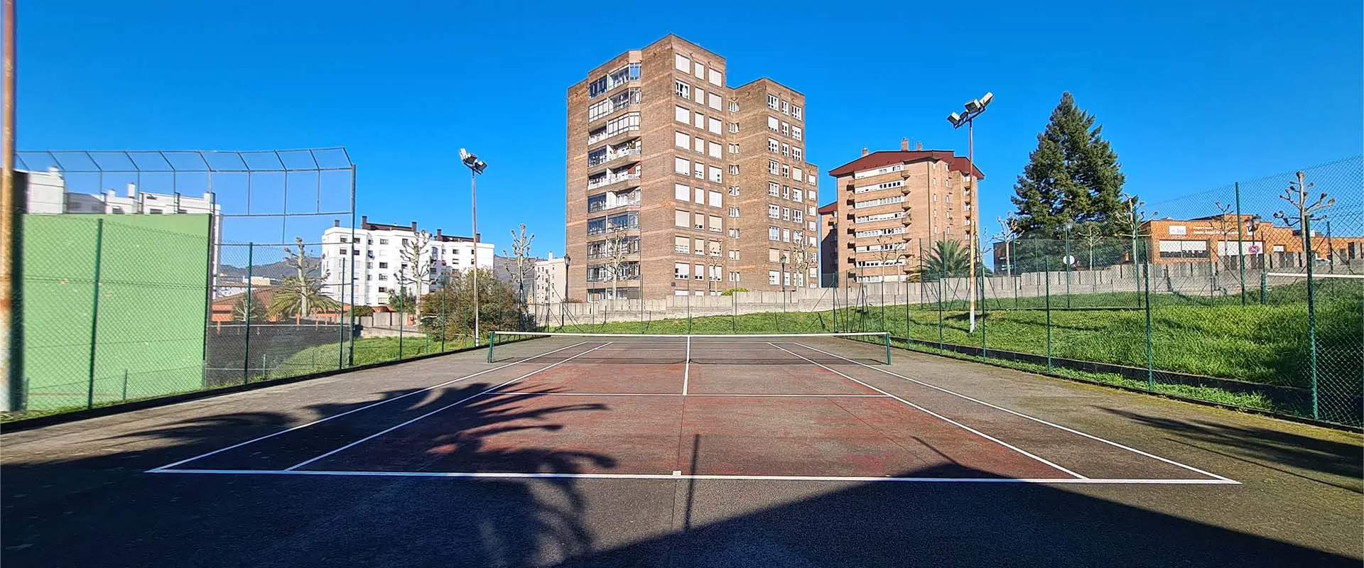 Vista frontal de una de las pistas de tenis, con el muro de cierre de las instalaciones y edificios circundantes al fondo