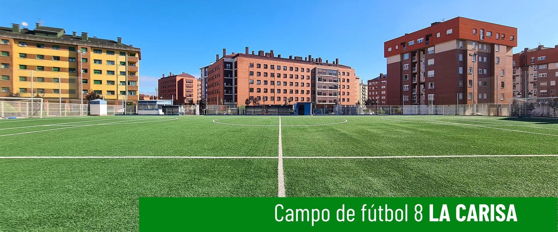 Foto del campo de fútbol 8 La Carisa.Terreno de juego a ras de césped. Líneas del campo en primer plano. Edificios al fondo 