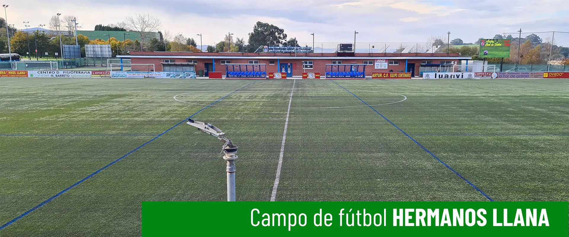 Imagen a ras de césped del campo de fútbol hermanos Llana. Cañón de riego en primer plano. Edificio de servicios al fondo 