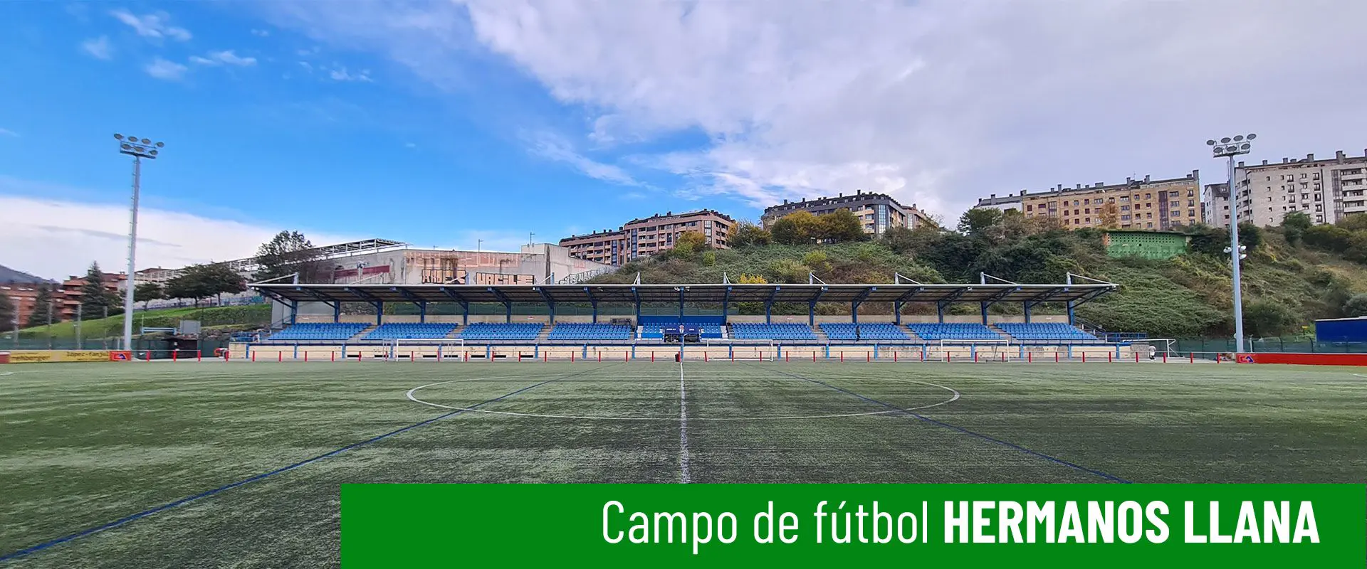 Campo de fútbol Hermanos Llana. Vista lateral del campo, con las gradas al fondo. Dos torres de iluminación a ambos lados 