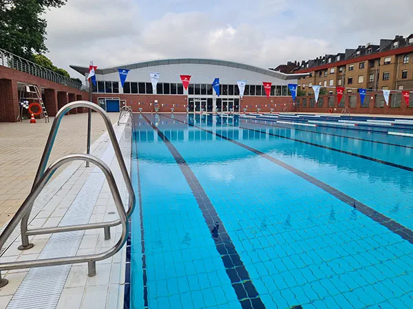 vista de la piscina desde una esquina, en primer plano escalerilla de acceso, al fondo edificio de piscinas cubiertas